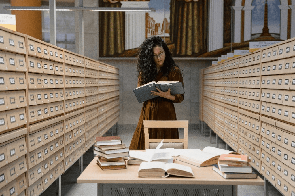 Researcher reading in a library aisle among wooden card catalogues and open books