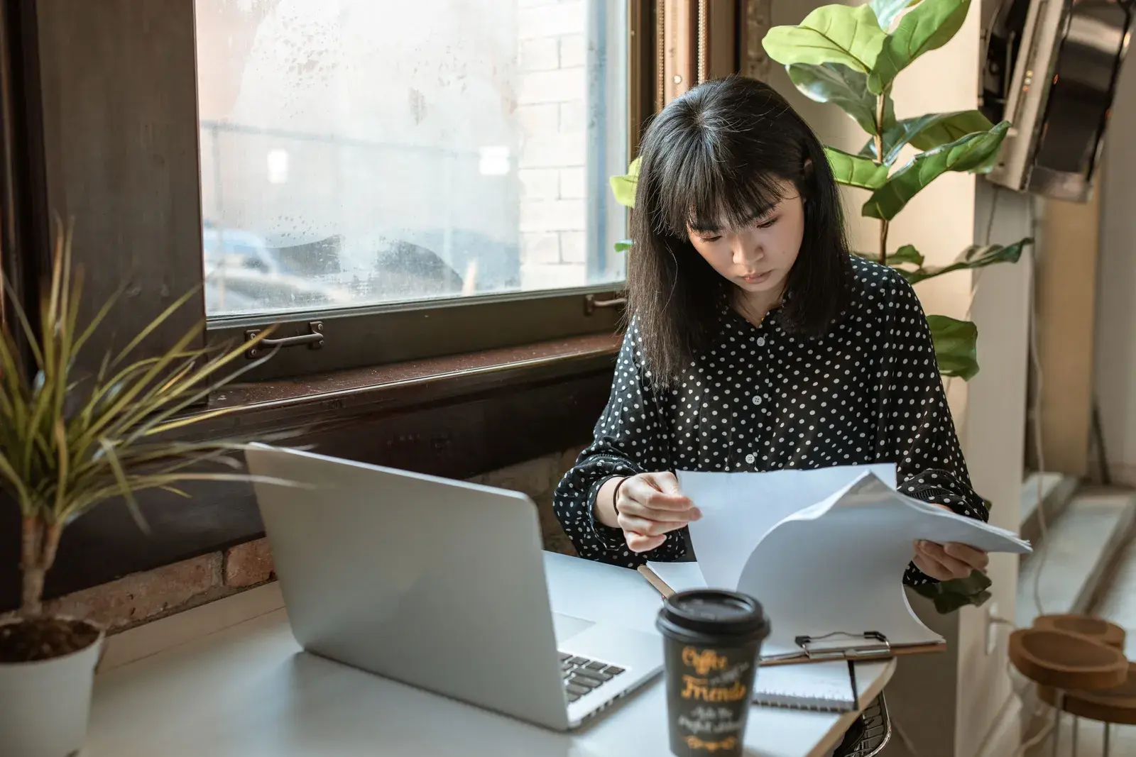 A woman reading through printed papers at a window table beside a laptop